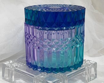 May include: A decorative jar with a faceted design, featuring a gradient of colors from blue to purple to teal. The jar sits on a clear, square, glass base. The lid is a deep blue color.