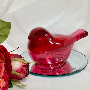 May include: A red glass bird figurine with a mirrored base. The bird is sitting on a white surface.