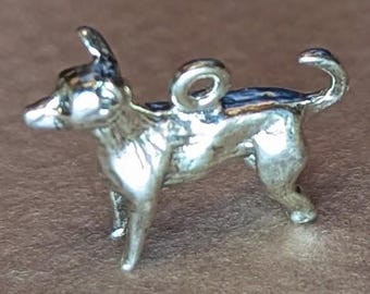 May include: A small, silver-colored charm in the shape of a dog, possibly a Chihuahua. The dog is standing with its tail curled up. It has a loop at the top for attaching to a bracelet or necklace. The dog's back has a dark blue accent.