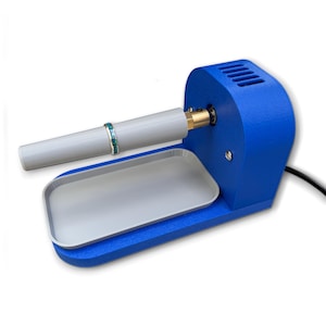 Può includere: Una lucidatrice per gioielli blu e grigia con un anello sull'asta di lucidatura. La macchina ha un vassoio grigio e una base blu. L'anello è argentato con dettagli blu e verdi. Un cavo di alimentazione nero esce dalla macchina.