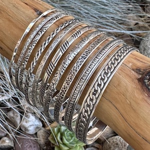 May include: A set of silver bangles with various embossed designs, including floral, geometric, and chain patterns. The bangles are stacked on a wooden surface.