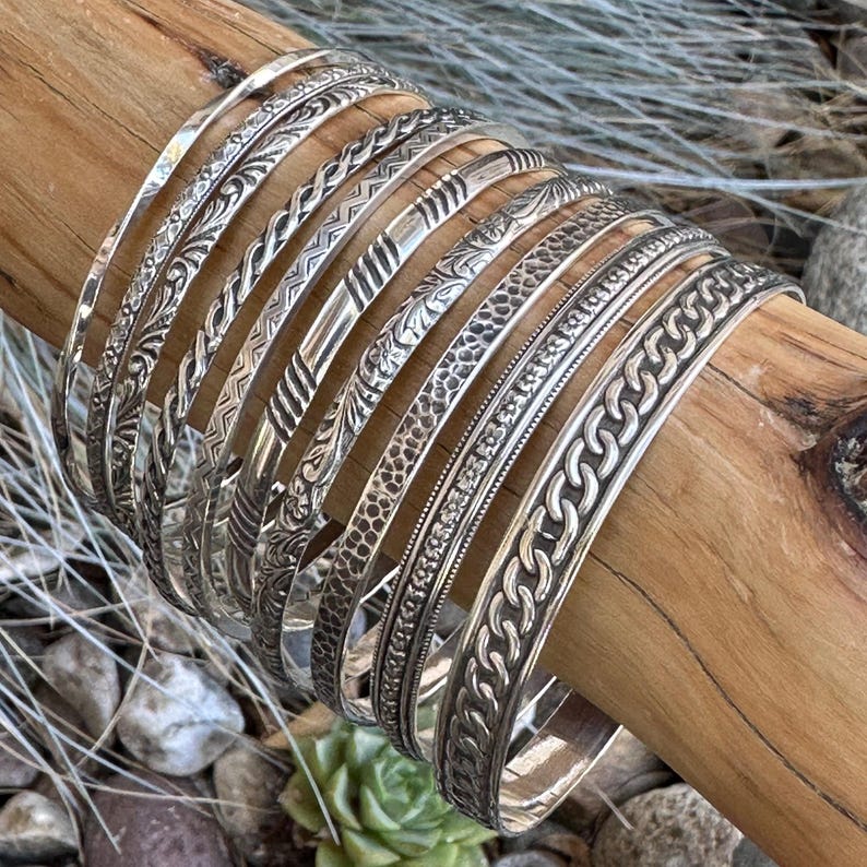 May include: A set of silver bangles with various embossed designs, including floral, geometric, and chain patterns. The bangles are stacked on a wooden surface.