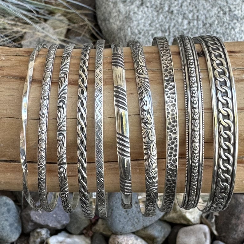 May include: A collection of silver bangles with various embossed designs, including floral, geometric, and braided patterns. The bangles are stacked on a wooden surface.