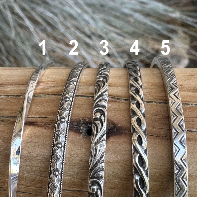 May include: Five silver bracelets with different embossed designs. The bracelets are arranged in a row on a wooden surface. The bracelets are numbered 1 through 5.