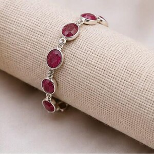May include: A silver chain bracelet featuring a series of oval-shaped, deep red ruby gemstones set in polished silver bezels. The bracelet consists of multiple linked segments, each holding a single faceted stone, creating a continuous design.