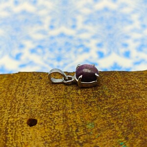 Puede incluir: Un colgante de plata con una piedra preciosa púrpura engastada en un engaste de plata. El colgante está sobre una superficie de madera.
