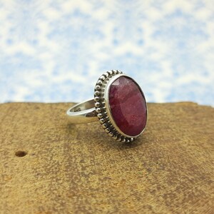 Può includere: Un anello d'argento con una grande pietra preziosa di rubino di forma ovale incastonata in una montatura a perline. L'anello è su una superficie di legno marrone.
