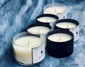 May include: Five black and clear glass candle jars with white labels. The labels have the text "The Crow's Magic" and "Intentional Divine Vibes" with a small illustration of a crow. The candles are on a blue and white textured surface.