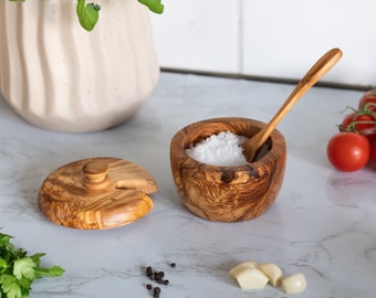 May include: A wooden salt cellar with a lid and a small wooden spoon. The cellar is filled with white salt and is sitting on a white marble surface. There are also some black peppercorns and garlic cloves on the surface.
