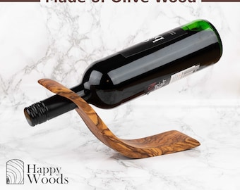 May include: A wooden wine bottle holder made of olive wood. The holder is shaped like a wave and is designed to hold a bottle of wine horizontally. The bottle holder is made of natural wood and has a rustic look. The bottle holder is a great way to display your favorite wine bottle.