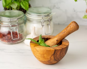 May include: A wooden mortar and pestle set with a green basil leaf inside. The mortar is made of olive wood and has a natural wood grain. The pestle is also made of olive wood and has a smooth, rounded handle.