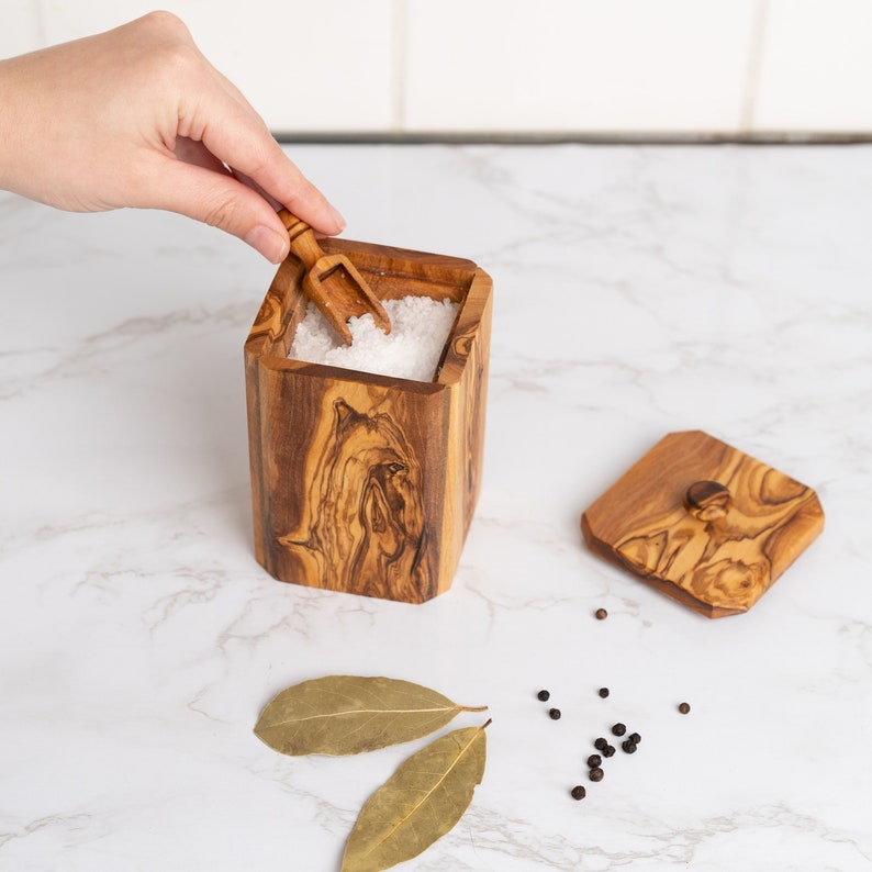 May include: A wooden salt cellar with a lid and a small wooden scoop. The cellar is filled with white salt and the scoop is resting on top of the salt. The cellar is made of olive wood and has a natural wood grain.