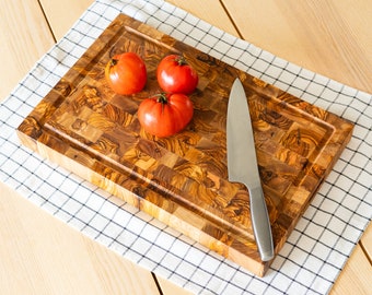 May include: A wooden butcher block cutting board with a juice groove, three red tomatoes, and a stainless steel chef's knife. The board has a mosaic pattern and is set on a checkered cloth.