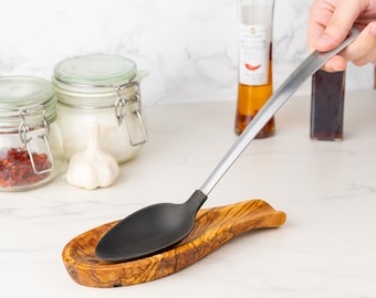 May include: A black cooking spoon rests in a wooden spoon rest. The spoon has a silver handle and a black head. The spoon rest is made of wood with a natural grain. Kitchen utensils.