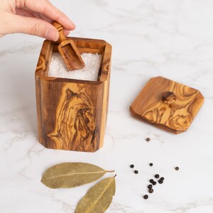 May include: A wooden salt cellar with a lid and a small wooden scoop. The cellar is filled with white salt. There are bay leaves and black peppercorns scattered on a white marble surface.