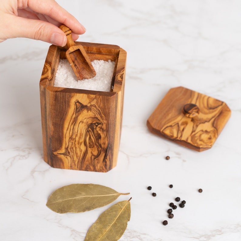 May include: A wooden salt cellar with a lid and a small wooden scoop. The cellar is filled with white salt. There are bay leaves and black peppercorns scattered on a white marble surface.