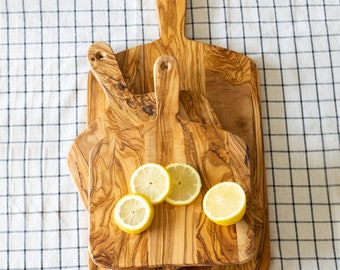 May include: Three wooden cutting boards with handles, stacked on top of each other. The boards are made of olive wood and have a natural, rustic finish. The top board has four lemon slices on it.