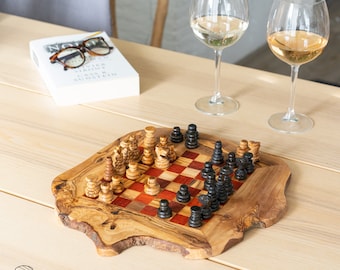 May include: A wooden chessboard with black and white squares and black and brown chess pieces. The board is made of olive wood and has a natural, rustic look. The chessboard is sitting on a wooden table with two wine glasses in the background.