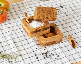 May include: A wooden salt and pepper shaker set with a lid and a small wooden spoon. The set is made of olive wood and has a natural finish. The shakers are filled with salt and pepper, and the spoon is resting on top of the salt shaker.