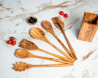 May include: A set of five olive wood kitchen utensils including a pasta fork, two spatulas, a slotted spoon, and a solid spoon, accompanied by a rectangular wooden utensil holder, arranged with small red tomatoes and a bowl of black olives.