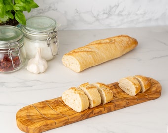 May include: A wooden bread board with grooves for slicing bread. The board is made of olive wood and has a natural finish. A baguette is sliced on the board, with the slices arranged in a row.