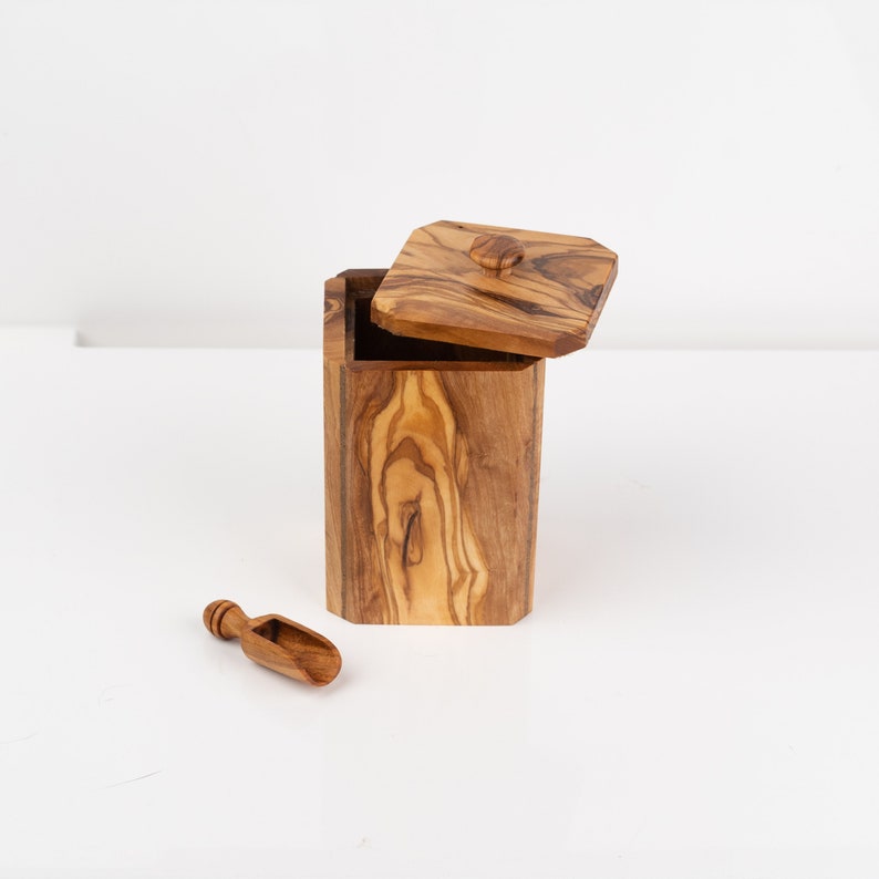 May include: A wooden box with a lid and a small wooden scoop. The box is made of olive wood and has a natural wood finish. The lid has a small knob on top. The scoop is about 3 inches long.