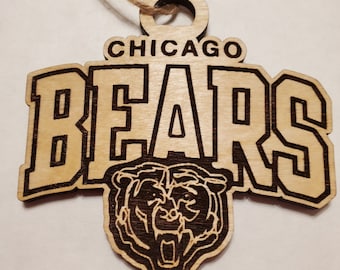 Może przedstawiać: Drewniana ozdoba Chicago Bears z nazwą drużyny i logo głowy niedźwiedzia. Ozdoba wykonana jest z jasnego drewna z ciemnobrązowymi wytrawionymi detalami i sznurkiem jutowym do zawieszenia.