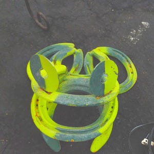 May include: A whimsical, handmade frog sculpture crafted from repurposed horseshoes. The sculpture features a vibrant color scheme of green and yellow paint, with the horseshoe shapes forming the frog's body and head. A unique piece of outdoor decor.