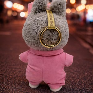 May include: A plush gray bunny toy wearing a pink jumpsuit and white shoes. The bunny has a gold-colored ring attached to its head with the text "THE MONSTER" printed on a gold strap. The background is a blurred street scene with bokeh lights.