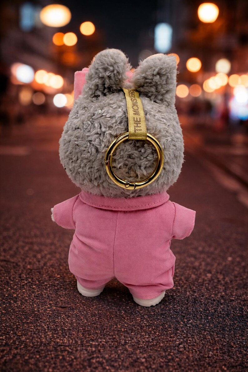 May include: A plush gray bunny toy wearing a pink jumpsuit and white shoes. The bunny has a gold-colored ring attached to its head with the text "THE MONSTER" printed on a gold strap. The background is a blurred street scene with bokeh lights.