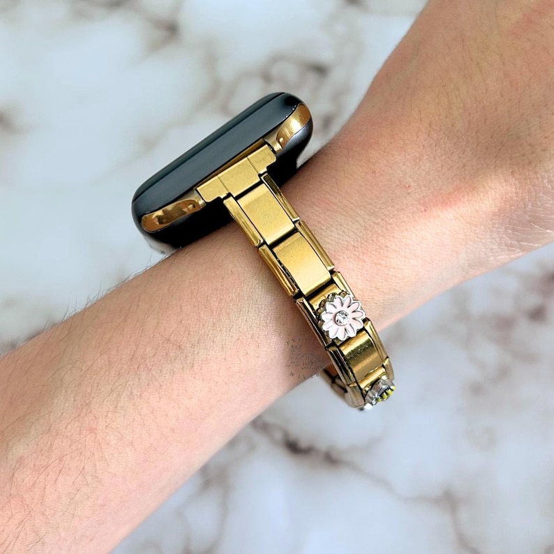 Op de afbeelding: Goudkleurige metalen horlogeband met een zwarte wijzerplaat en gouden rand. De band heeft een decoratieve bloem met roze bloemblaadjes en kleine heldere kristallen. Het horloge wordt om de pols gedragen.