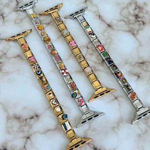 Op de afbeelding: Vier horlogebandjes met decoratieve bedels. Twee zijn goudkleurig en twee zilverkleurig. Elke band heeft een reeks kleine, kleurrijke bedels met verschillende ontwerpen, waaronder bloemen, smiley-gezichten en geometrische vormen. De bandjes worden weergegeven op een marmeren oppervlak.