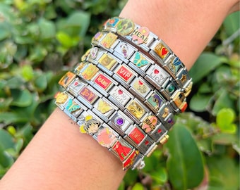 Op de afbeelding: Een stapel zilverkleurige schakelarmbanden met diverse kleurrijke emaille bedels, waaronder vlinders, harten, sterren en teksten zoals HEAL, VIP, I Love My Life, TeAmo, I Love My Girls, MOMMY'S GIRL, HEALTH en AMOR, met diverse edelsteenaccenten.