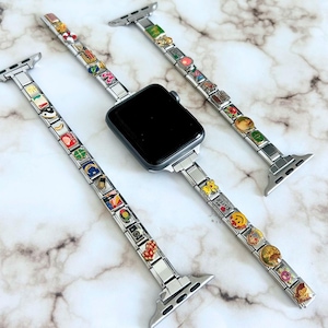 Op de afbeelding: Een zilveren Apple Watch met drie verwisselbare bandjes. Elke band is versierd met kleurrijke bedels. De bedels hebben verschillende ontwerpen, waaronder bloemen en smiley-gezichten. Het horloge en de bandjes worden gepresenteerd op een marmeren oppervlak.