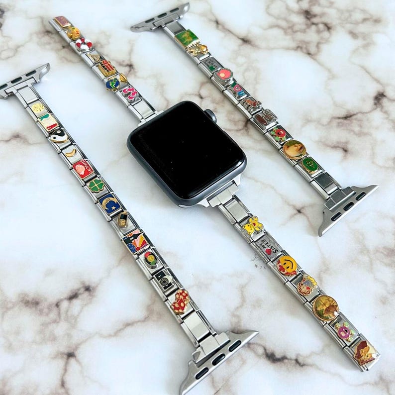Op de afbeelding: Een zilveren Apple Watch met drie verwisselbare bandjes. Elke band is versierd met kleurrijke bedels. De bedels hebben verschillende ontwerpen, waaronder bloemen en smiley-gezichten. Het horloge en de bandjes worden gepresenteerd op een marmeren oppervlak.