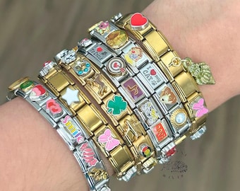Op de afbeelding: Een collectie van modulaire schakelarmbanden in goud- en zilverkleur met diverse decoratieve bedels, waaronder hartjes, een klavertje, een kat, een fiets, een vliegtuig, een parel en teksten zoals I LOVE TO CYCLE en I LOVE MY CAT met schelpbedel.
