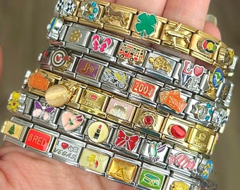 Op de afbeelding: Een verzameling bedelarmbanden met diverse decoratieve bedels. De bedels bevatten een klavertjevier, een Londense bus, een hart, een lippenstift en het nummer "2003". De armbanden zijn gemaakt van goud- en zilverkleurig metaal.