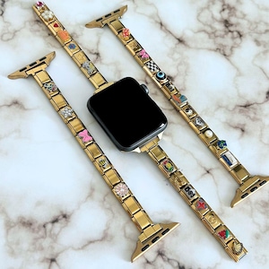 Op de afbeelding: Goudkleurige Apple Watch-bandjes met kleurrijke bedels. De bandjes zijn versierd met diverse decoratieve bedels in verschillende vormen en kleuren. De wijzerplaat van de horloge is zwart. De bandjes worden gepresenteerd op een marmeren oppervlak.