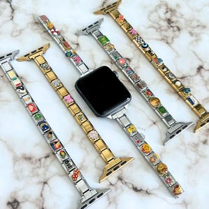 Puede incluir: Una variedad de correas para Apple Watch con dijes decorativos. Las correas son plateadas y doradas, con varios dijes que incluyen flores, caras sonrientes y formas geométricas. Un Apple Watch negro está en el centro, sujeto a una correa plateada.