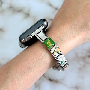 Op de afbeelding: Zilveren horlogeband met decoratieve bedels. De bedels omvatten een groene vierkant, een goudkleurige bedel met een kroon en een klaver, en een witte bedel met tekst. De wijzerplaat van het horloge is zwart.