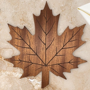 May include: A dark brown wooden cutting board shaped like a large maple leaf. The board features a smooth surface with dark, etched lines that form the intricate vein pattern of a leaf, extending from the central stem to the pointed edges.