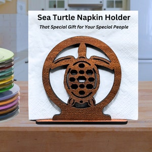 May include: A wooden sea turtle napkin holder with the text "Sea Turtle Napkin Holder" and "That Special Gift for Your Special People". The holder is brown and holds white napkins. A stack of colorful plates and bowls are in the background.