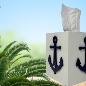 May include: White tissue box cover with two blue anchor designs. The tissue box is on a wooden surface with a palm tree in the background.