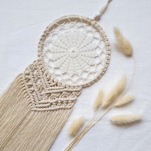 Può includere: Un acchiappasogni in macramè beige con un centro in crochet bianco e una lunga frangia. L'acchiappasogni è appeso a una superficie bianca con erba della pampa secca.