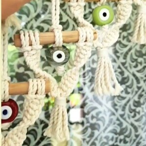 May include: A white macrame wall hanging with wooden dowels and blue, green, and red evil eye beads.