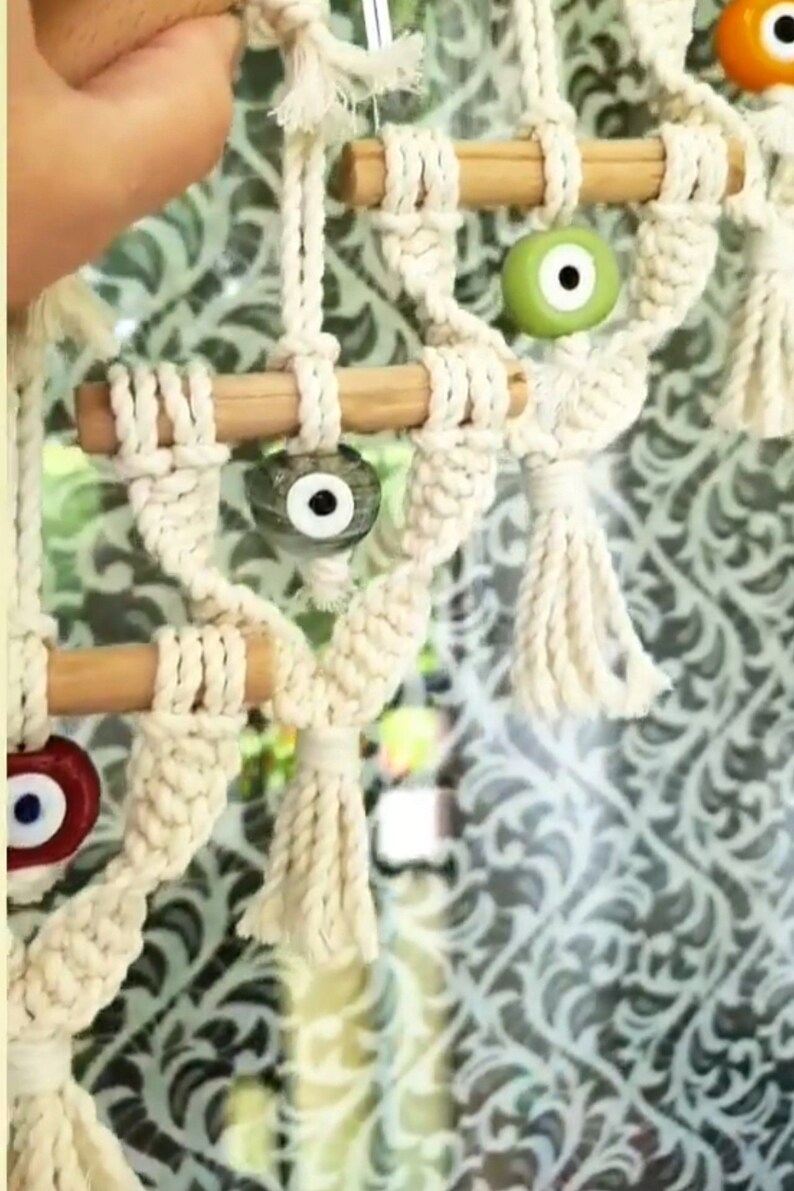 May include: A white macrame wall hanging with wooden dowels and blue, green, and red evil eye beads.