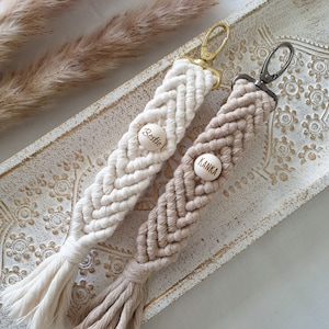 Può includere: Due portachiavi in macramè bianco e beige con perline di legno. Le perline sono incise con le parole "Bestie" e "Kank".