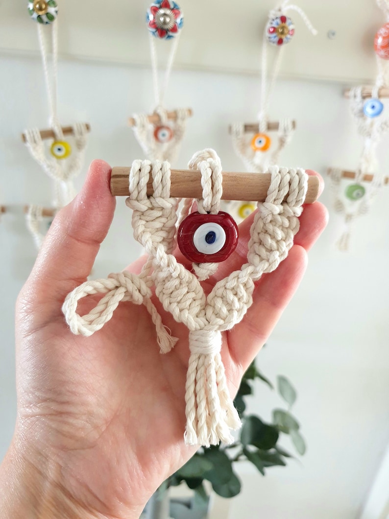 May include: A small macrame wall hanging with a red evil eye charm. The hanging is made with white cotton cord and has a wooden dowel at the top.