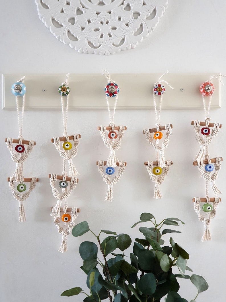 May include: Five white macrame wall hangings with blue, green and orange evil eye beads. Each hanging has three tiers and is attached to a knob on a white wall.