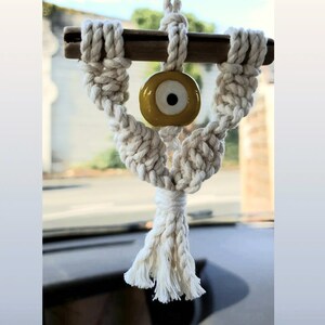 May include: White macrame car charm with a yellow evil eye bead. The charm is hanging from a rearview mirror.